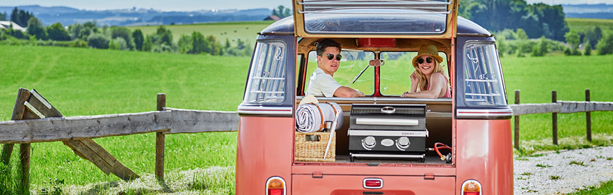 Gasgrill BBQ-Portable Videro G2-P Paar schaut aus einem VW Bus mit Gasgrill BBQ-Portable Videro G2-P im Kofferraum